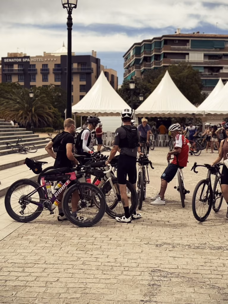 Riders with loaded gravel bikes standing in a paved square in front of white event tents.