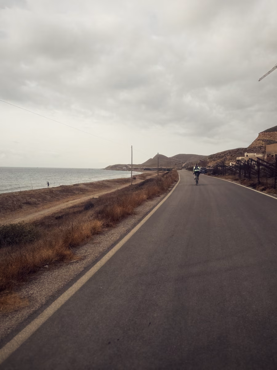 A coast road under a gray sky, the sea on the left, a lone rider ahead, dry hills on the right.