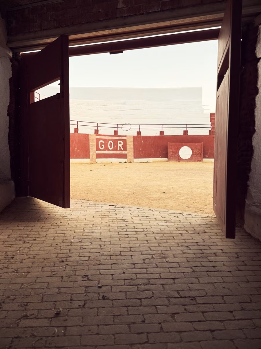 The open wooden gate of a small bullring, GOR painted in white on the red barrier inside, empty sand and white stands beyond.