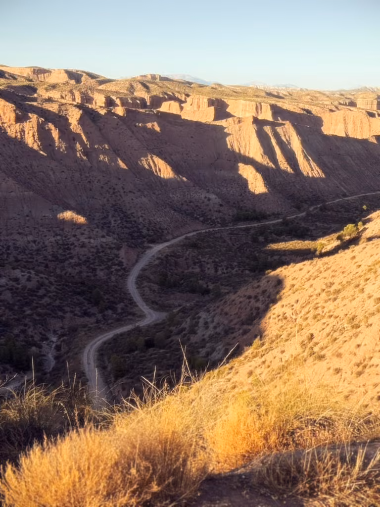 A dirt road curves through a deeply eroded canyon, with sunlit slopes above and long shadows below.