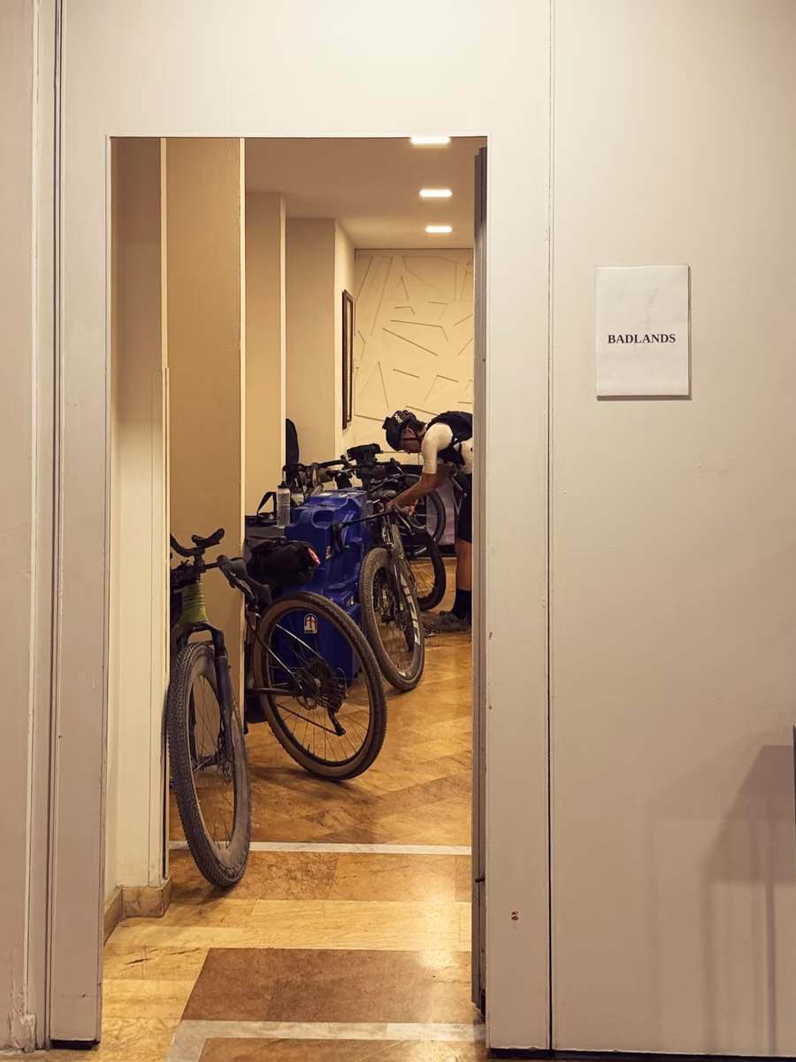 Loaded gravel bikes line a room off a hotel hallway. A paper sign on the door reads BADLANDS.