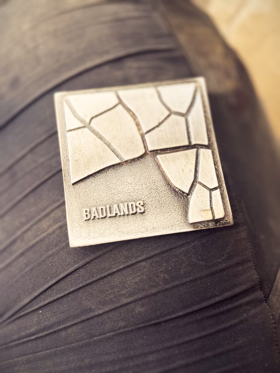 A square metal finisher's medal with a cracked-clay relief and the word BADLANDS, resting on a rider's thigh.