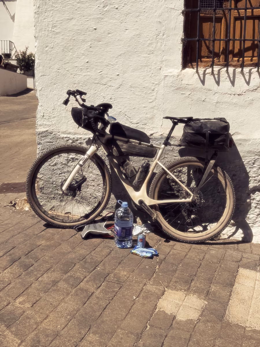 A loaded gravel bike leans against a whitewashed wall, with water bottles on the pavement.