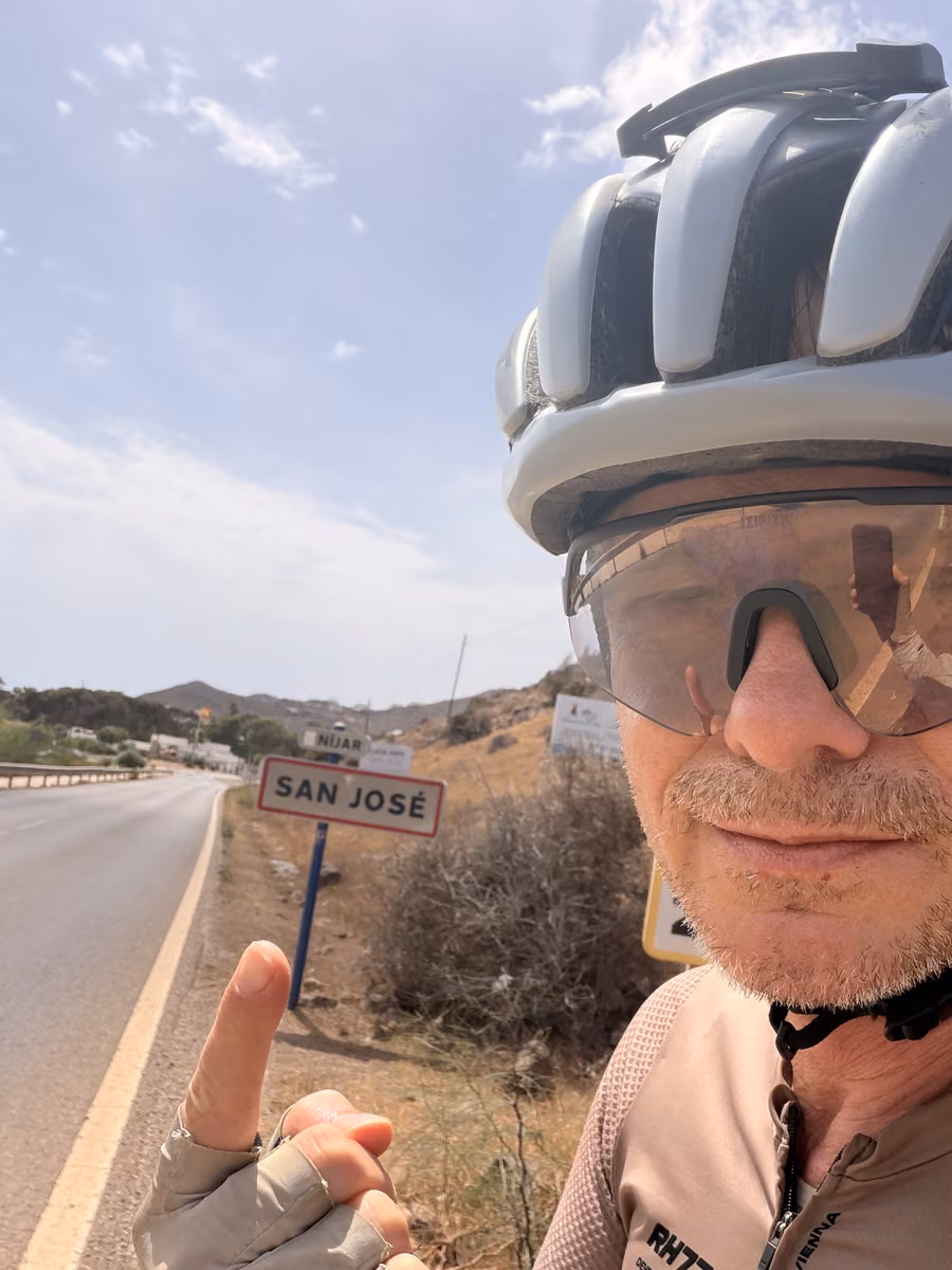 Selfie in helmet and sunglasses pointing at the San José village sign beside the road.