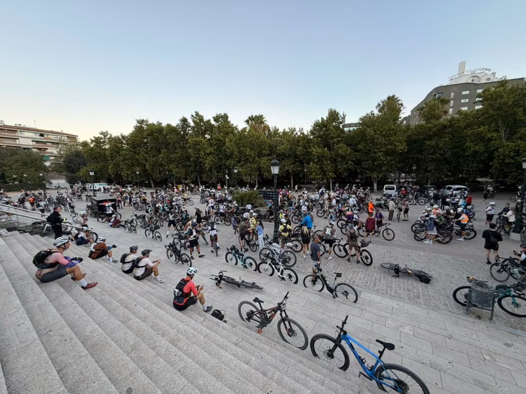 Riders and bikes fill the square below a wide flight of steps, with people waiting along the edges.