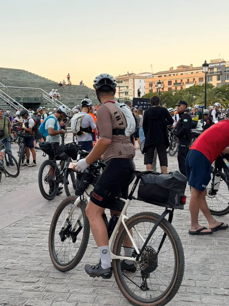 A rider stands with a loaded gravel bike among people gathering beside broad stone steps.
