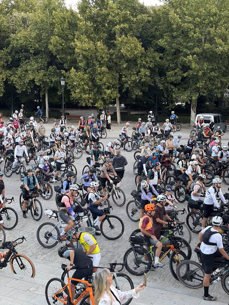 Several hundred riders with loaded gravel bikes waiting under plane trees in a Granada square, seen from above.