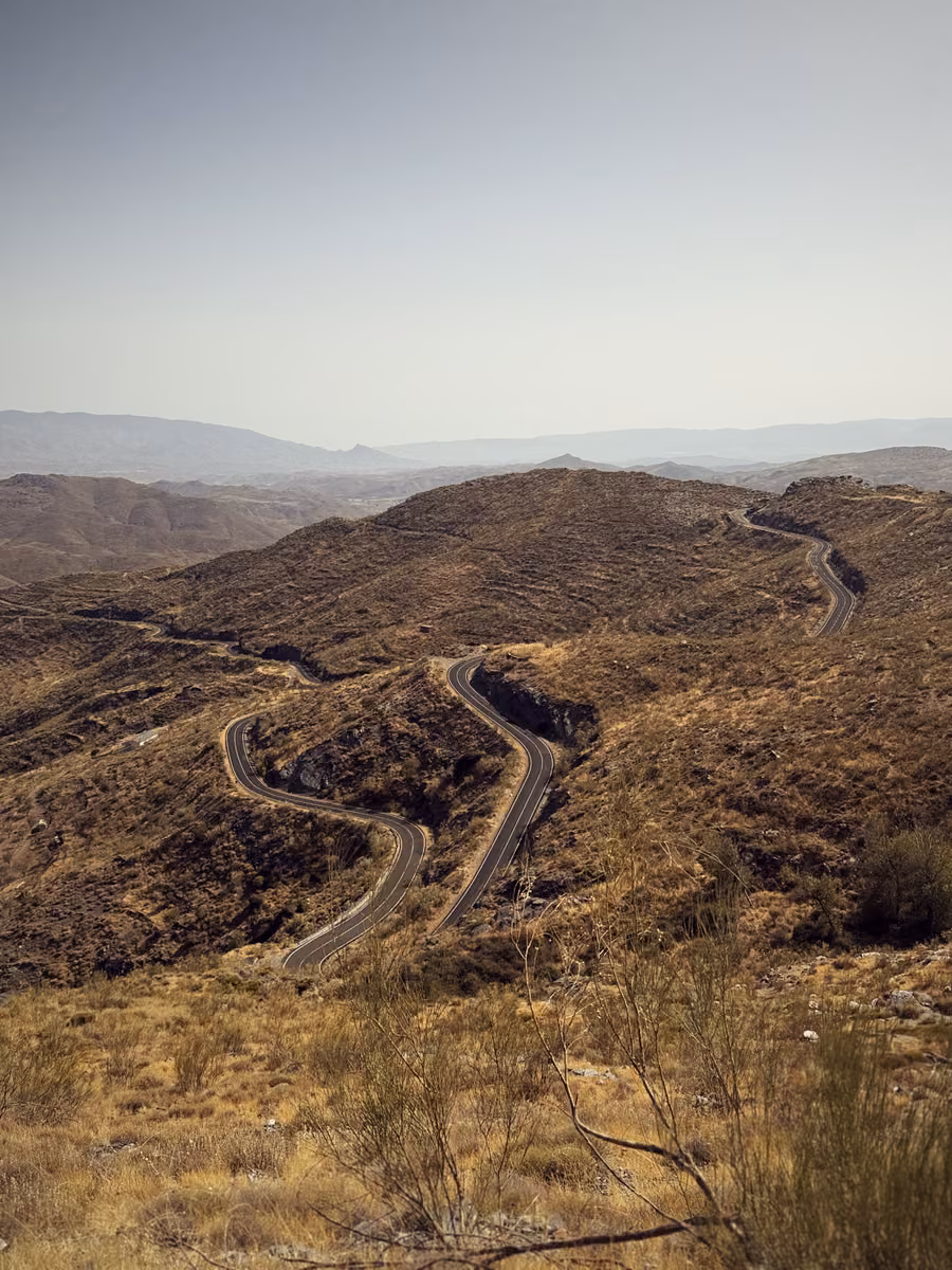 A narrow road switchbacking down brown mountainsides toward a hazy plain.